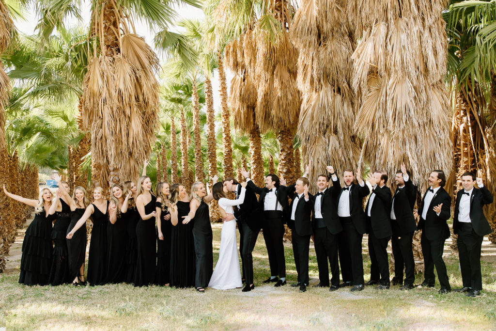 bride and groom kiss while bridal party of nine on each side all in black cheers