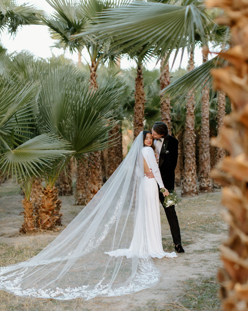 boujee wedding couple poses in palm tree oasis. groom kisses bride on forehead from the side.