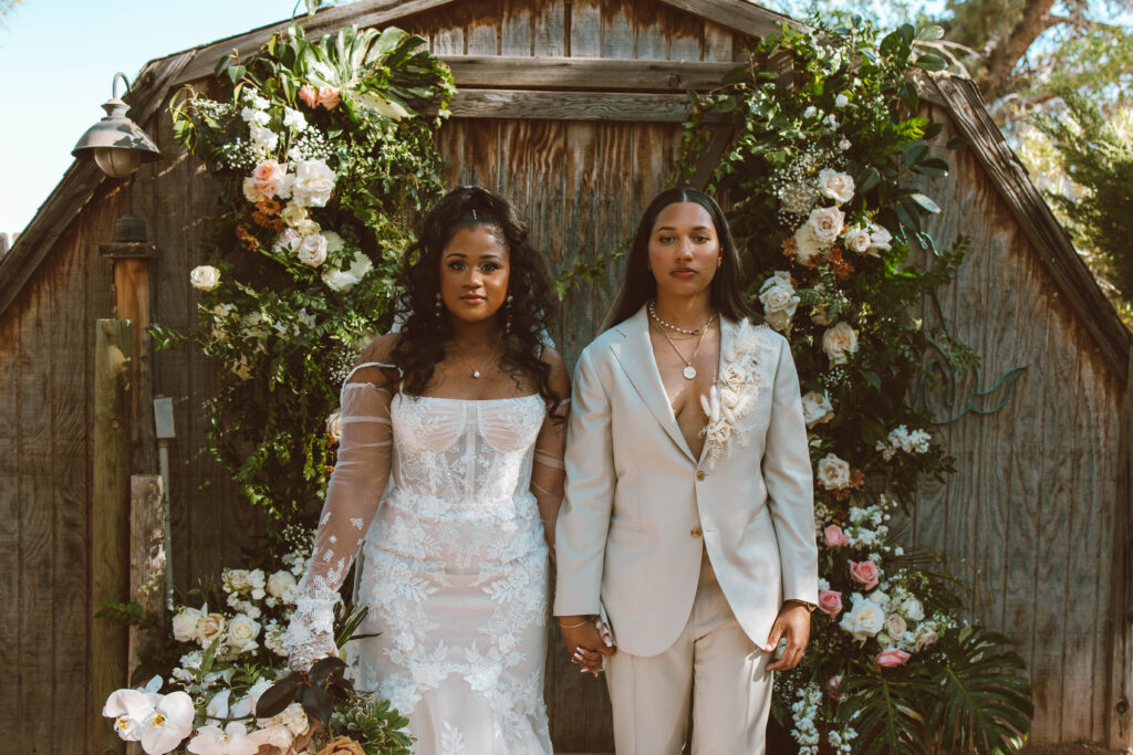 modern, boho glamour shot of both brides standing side by side in front of barn doors decorated with garden chic flower arrangements 
