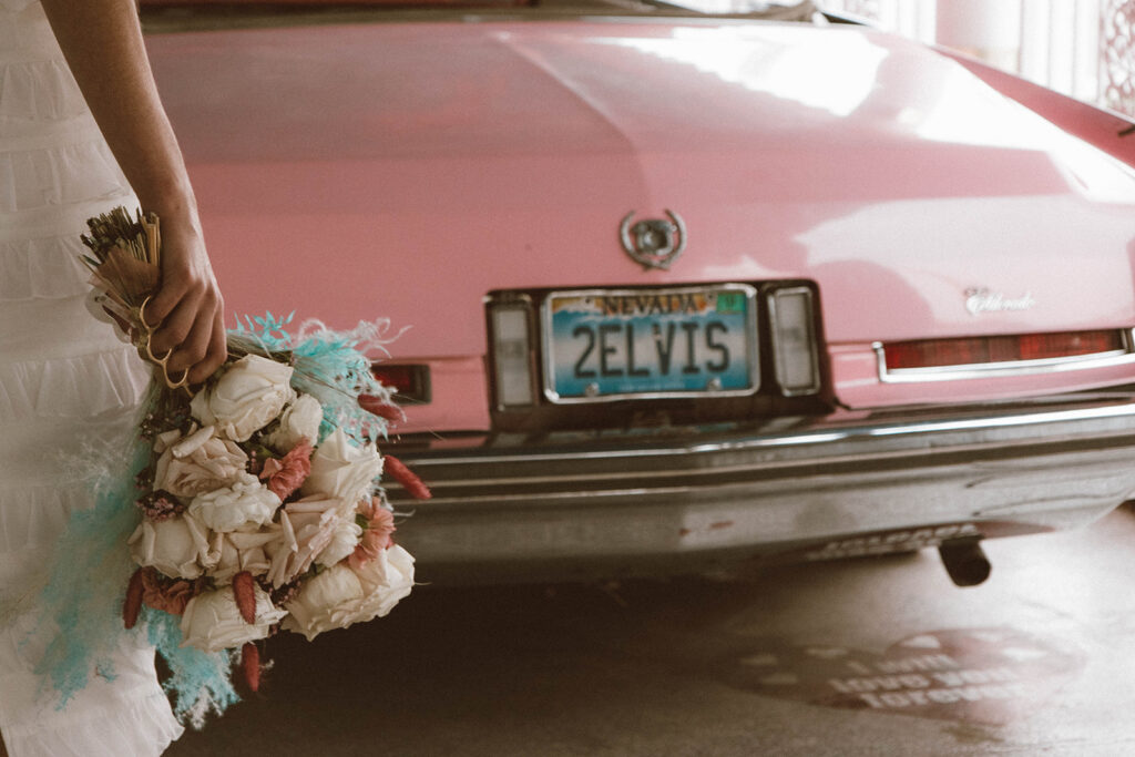 Bride holding a colorful bouquet beside the pink Cadillac at A Little White Wedding Chapel in Las Vegas